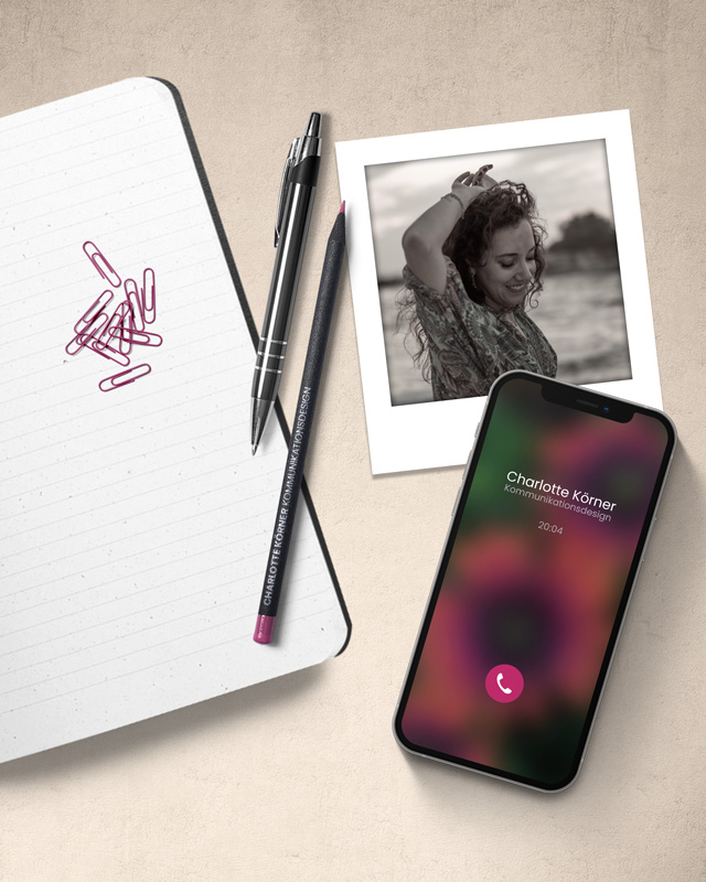 charlotte-grafikdesign Notizbuch, Stifte, Büroklammern. Ein Handy, auf dem Gerade jmd. anruft. Ein Foto von Charlotte Körner als Polaroid. Die ganze Szene ist von oben aufgenommen worden.